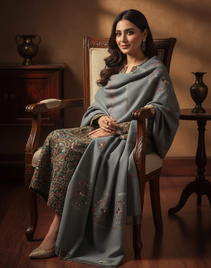 Woman sitting on a wooden chair wearing a gray shawl in a warm-toned room.